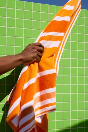 Close-up of a dark-skinned arm holding an orange and white striped towel against a bright green tiled wall in sunlight.