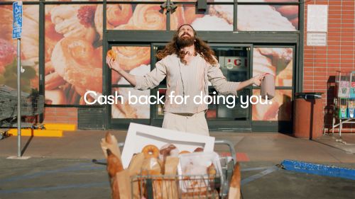 Adult male with long hair stands outside a bakery with a shopping cart full of bread and a donut in his hand, advertising cash back.