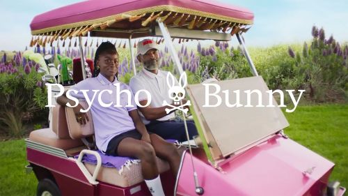 Two people in a pink golf cart on a golf course with purple flowers and greenery. Polo shirts, hats, skull logo, and a sunny day.