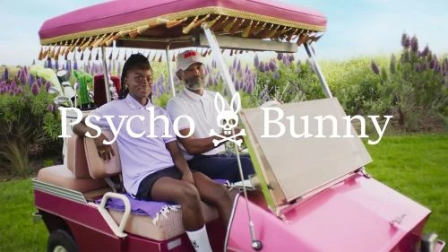 Two people in a pink golf cart on a golf course with purple flowers and greenery. Polo shirts, hats, skull logo, and a sunny day.