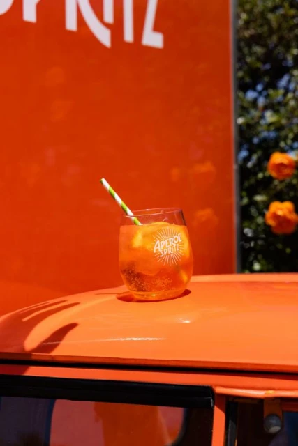 Aperol spritz in glass with straw on a bright orange surface with white brand text, capturing a refreshing and vibrant beverage presentation.