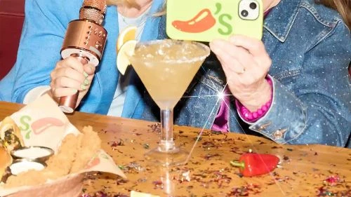 Two people pose at a restaurant table with cocktails, a microphone, and appetizer plates, featuring a red pepper prop and colorful confetti.