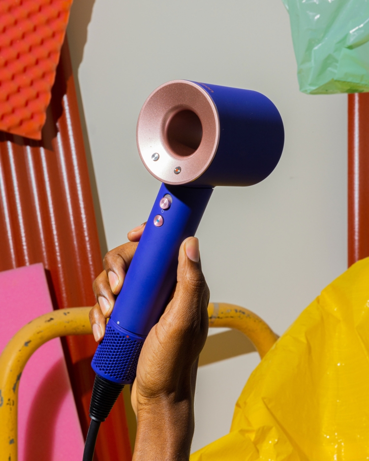 Hand holding a blue and rose gold Dyson hair dryer in front of colorful panels and plastic bags, indoor product shot, beauty product.