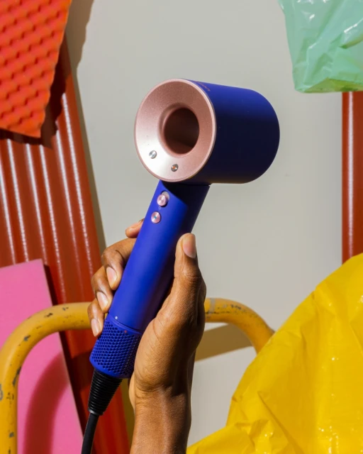Hand holding a blue and rose gold Dyson hair dryer in front of colorful panels and plastic bags, indoor product shot, beauty product.