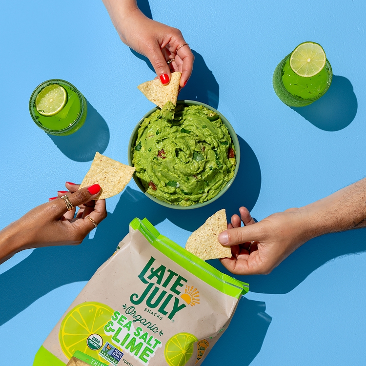 Overhead shot of Late July organic sea salt and lime tortilla chips, guacamole, and drinks with lime slices, hands dipping chips.