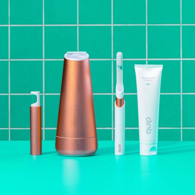 Dental hygiene products including an electric toothbrush, toothpaste and charger on a bright turquoise surface against a green tiled wall.