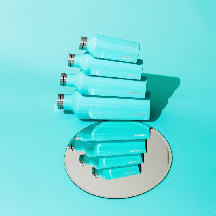 Still life studio shot of a stack of four matching teal water bottles and their reflections on a round mirror over a teal background.