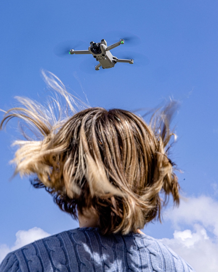 A drone flies above a person with wavy blonde hair outside, with a clear blue sky and scattered clouds in the background.