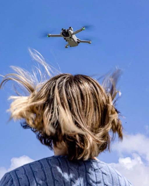 A drone flies above a person with wavy blonde hair outside, with a clear blue sky and scattered clouds in the background.
