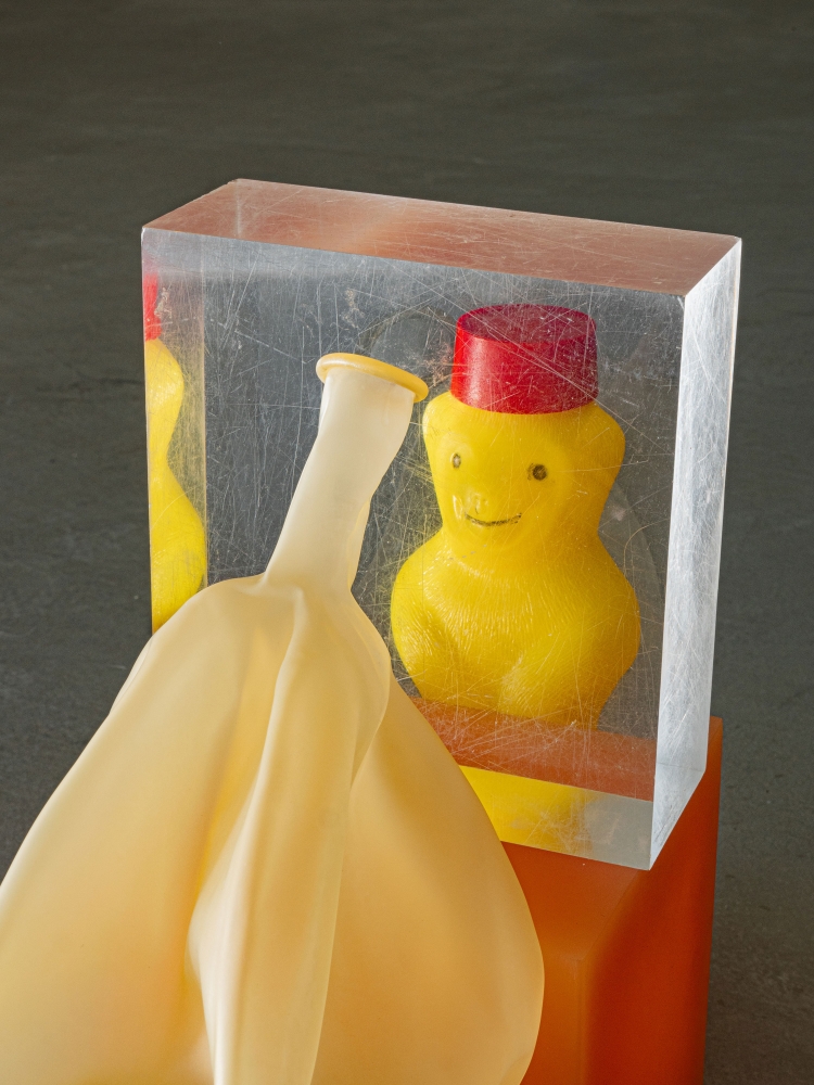 A yellow toy duck with a red hat encased in a clear block sits next to a deflated balloon on a gray surface with an orange base.