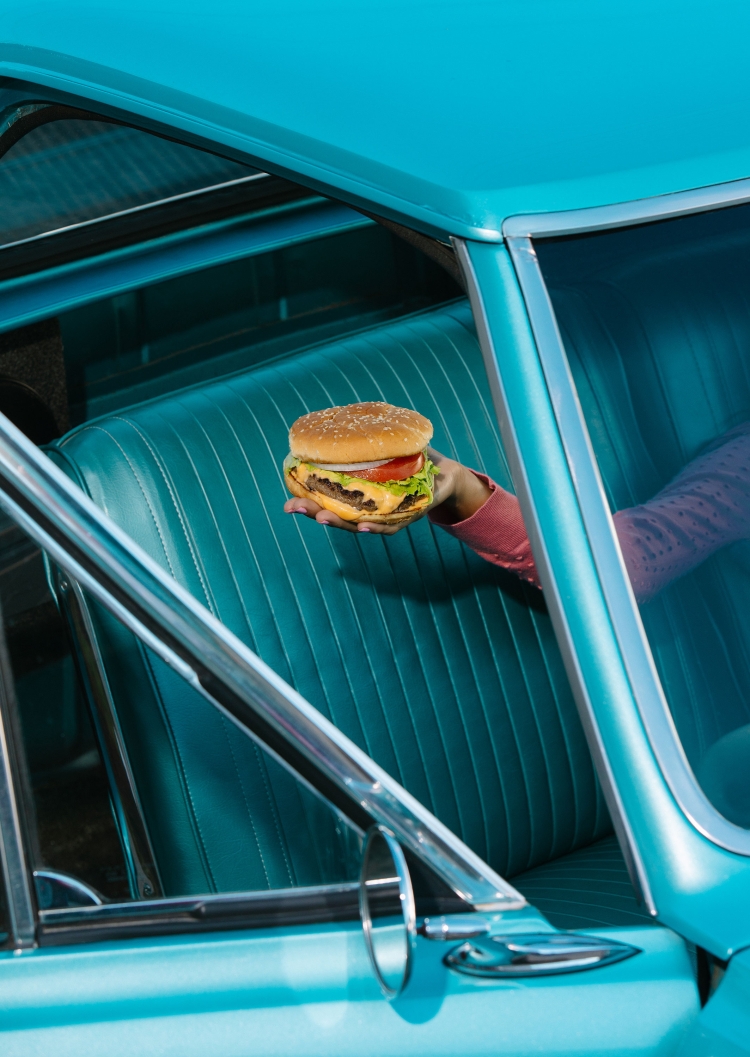 Close-up of a hand holding a burger in a car with turquoise interior and exterior, capturing a vintage aesthetic and food presentation.