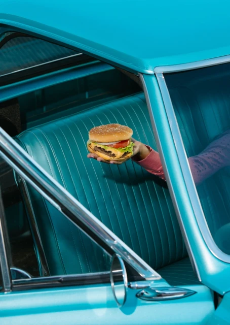 Close-up of a hand holding a burger in a car with turquoise interior and exterior, capturing a vintage aesthetic and food presentation.