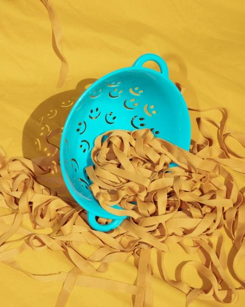 Overhead studio shot of a blue smiley-face colander with dried tan-colored pasta spilling out onto a solid mustard-yellow backdrop.
