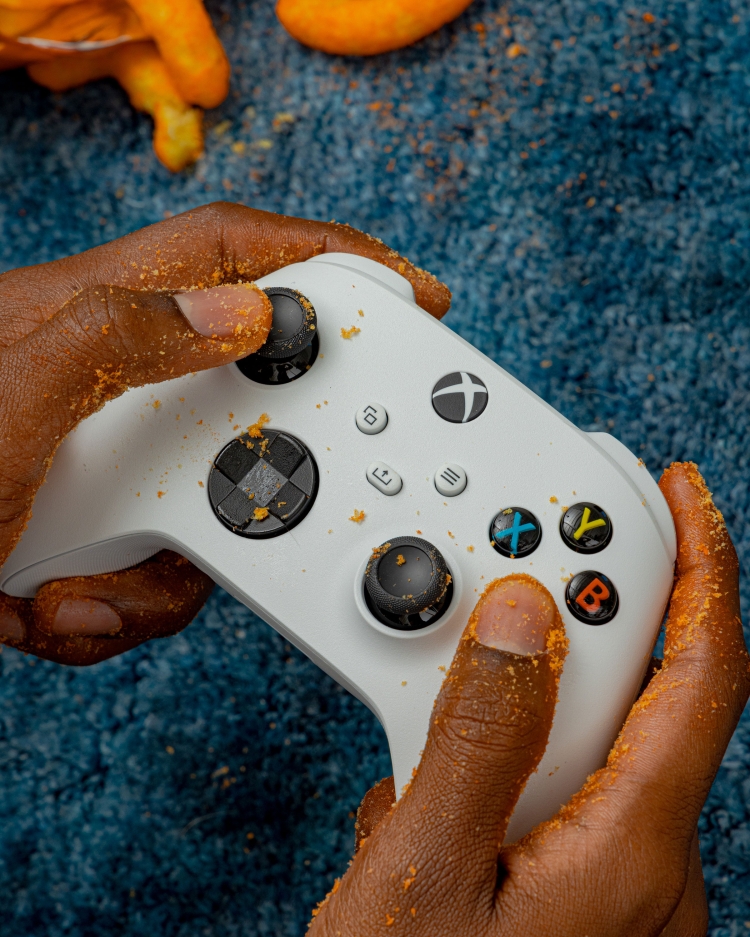 Close-up of dark-skinned adult hands holding a white Xbox controller with snack dust, set against a textured blue background.