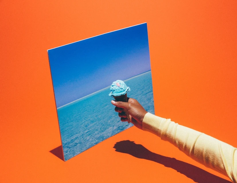 Hand holding blue ice cream cone against a backdrop showing the ocean and sky, with a bright orange background creating a vibrant, summery image.