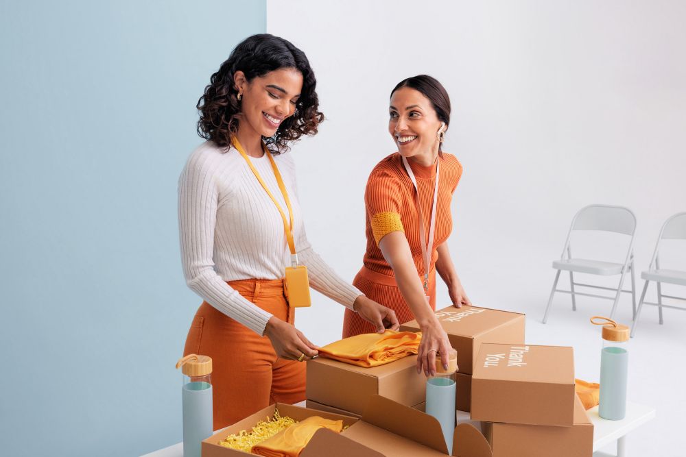 Two smiling women pack boxes with orange t-shirts and water bottles, standing at a white table against a split blue-and-white background.