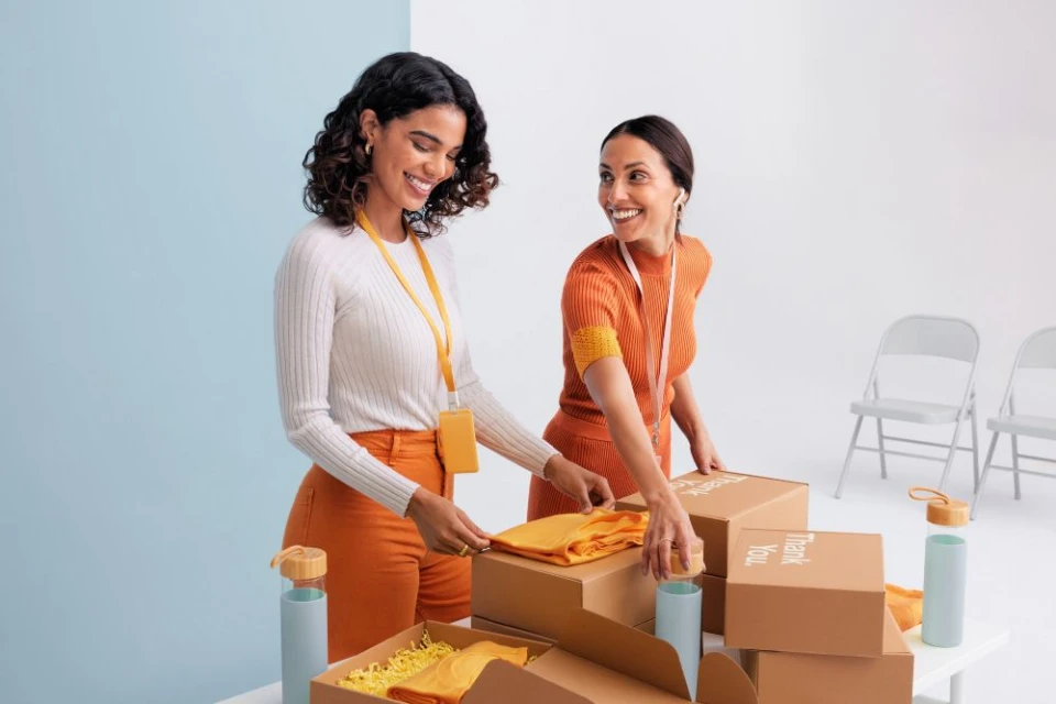 Two smiling women pack boxes with orange t-shirts and water bottles, standing at a white table against a split blue-and-white background.