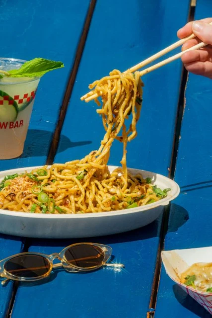 Close-up of noodles in a white bowl being lifted with chopsticks on a blue table next to a drink and sunglasses.