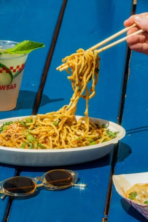 Close-up of noodles in a white bowl being lifted with chopsticks on a blue table next to a drink and sunglasses.