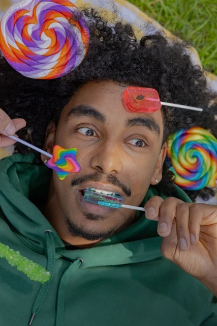 Person lying on grass surrounded by lollipops, wearing a green hoodie, posing with colorful star, swirl, and disc-shaped lollipops.