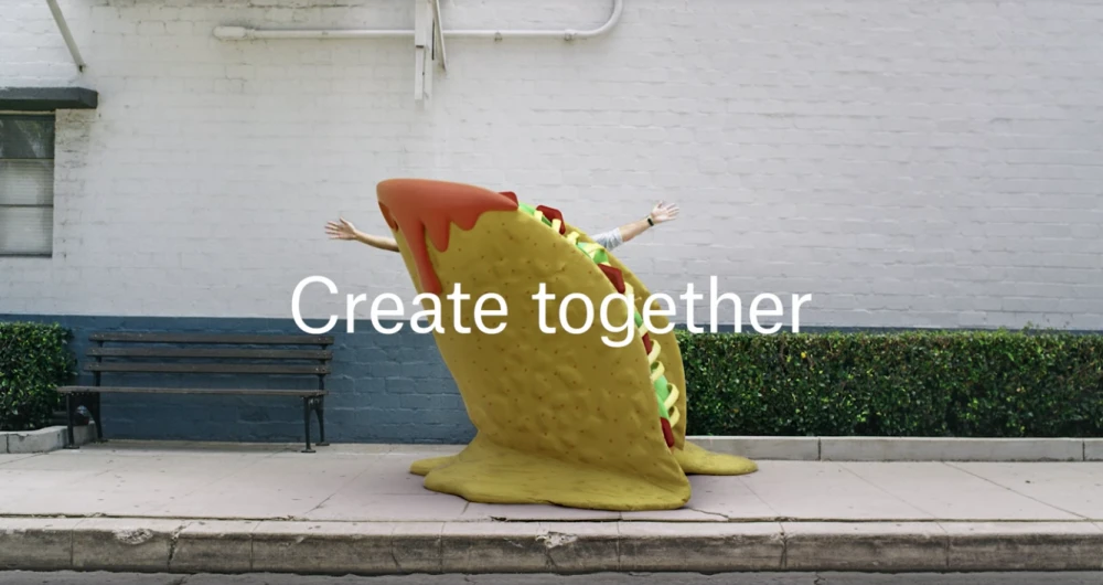 Person inside a large taco prop against a white brick building with green hedge. Conceptual visual content creation and food advertisement.