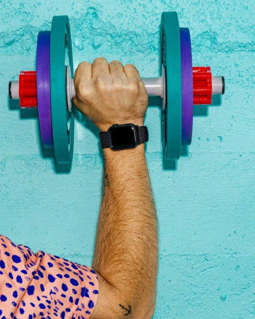 Person holding colorful dumbbell. Focus on hand, wrist with smart watch, arm, and part of the torso against blue textured background.