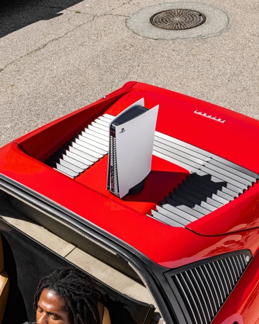 Overhead shot of a white PlayStation 5 on the hood of a red Ferrari, next to a person partially visible at the bottom.