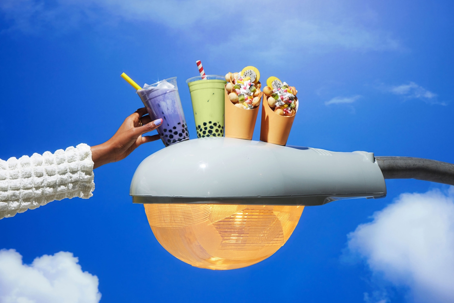 Hand presents bubble tea and waffle cone desserts on a light post against blue sky. Food photography highlighting sweet treats.