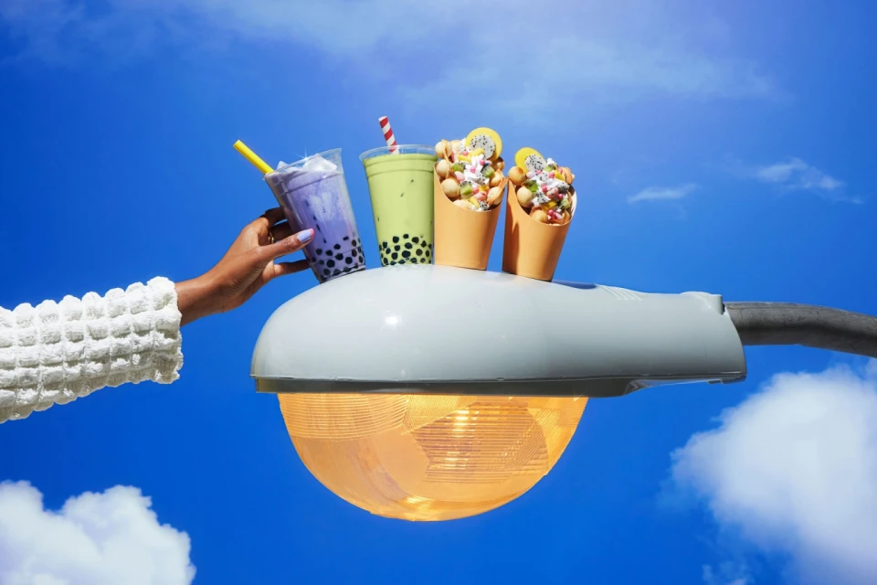 Hand presents bubble tea and waffle cone desserts on a light post against blue sky. Food photography highlighting sweet treats.