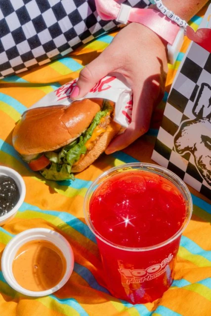 Eye-level shot of a hand holding a vegan burger, with a red beverage and sauces on a patterned surface, promoting food and beverage.