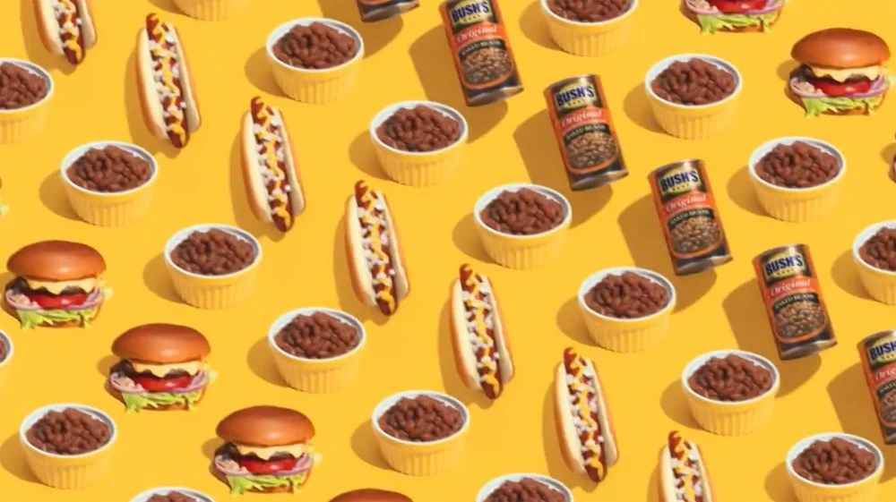 Overhead pattern on yellow backdrop: canned beans, bowls of baked beans, hot dogs with mustard, and cheeseburgers with lettuce and tomato.