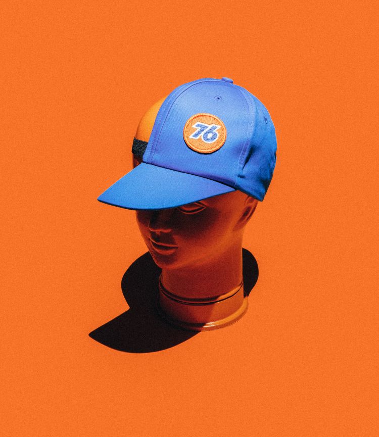Blue baseball cap with a '76' logo sits atop a mannequin head against a vibrant orange background, showcasing a blend of sporty style.