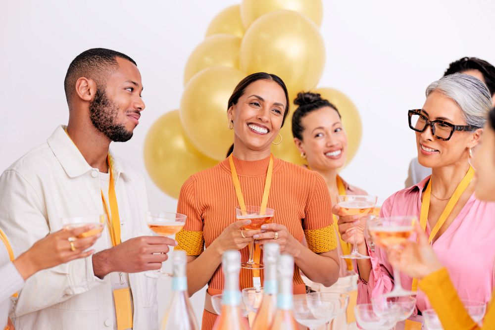 A group of six smiling adults in bright, casual clothing holding drinks at an event, with gold balloons and bottles in the background.