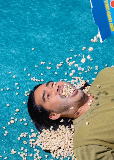 Adult male lying on blue surface with cereal box pouring cereal into his mouth, open mouth expression, sunny day, casual clothing.