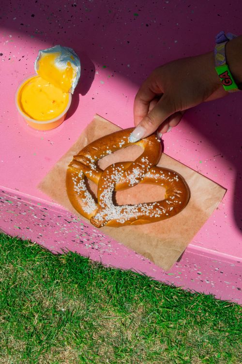 Overhead shot of a pretzel on brown paper with cheese dip, set on a pink speckled table with green grass visible. A hand reaches for it.