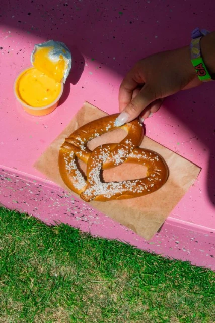 Overhead shot of a pretzel on brown paper with cheese dip, set on a pink speckled table with green grass visible. A hand reaches for it.
