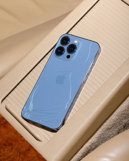 Light blue iPhone 13 Pro Max with a cracked screen is displayed on a beige leather car console in a studio product shot.