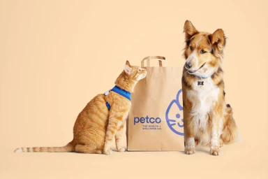 An orange tabby cat and a fluffy brown and white dog sitting beside a branded Petco paper shopping bag on a beige background.