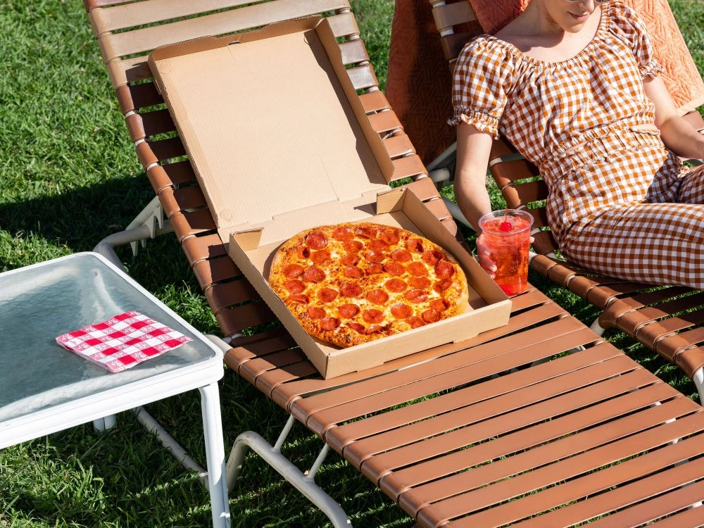 An open pizza box with a pepperoni pizza rests on a brown lounger beside a person wearing a checkered outfit.