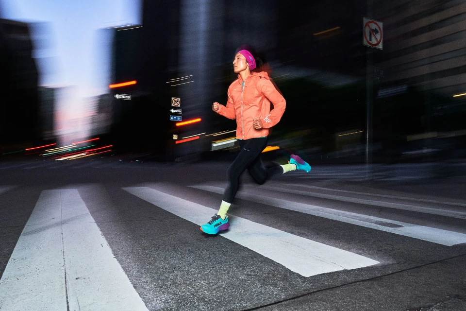 Female runner in an orange jacket runs across a white crosswalk on a dark city street with blurred lights and buildings at night.