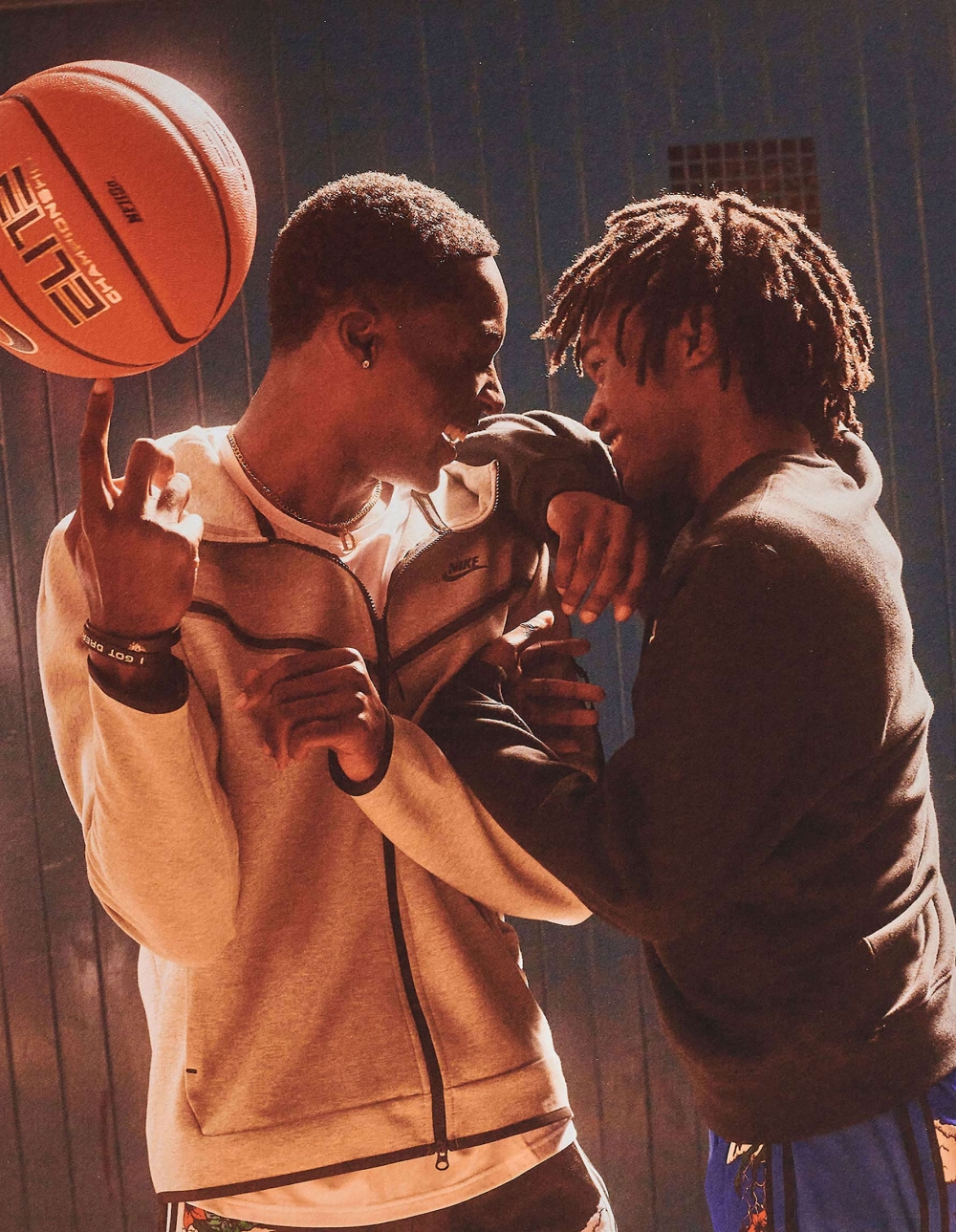 Two young men indoors: one spins a basketball on his finger while the other rests his arm on his shoulder in a friendly gesture.