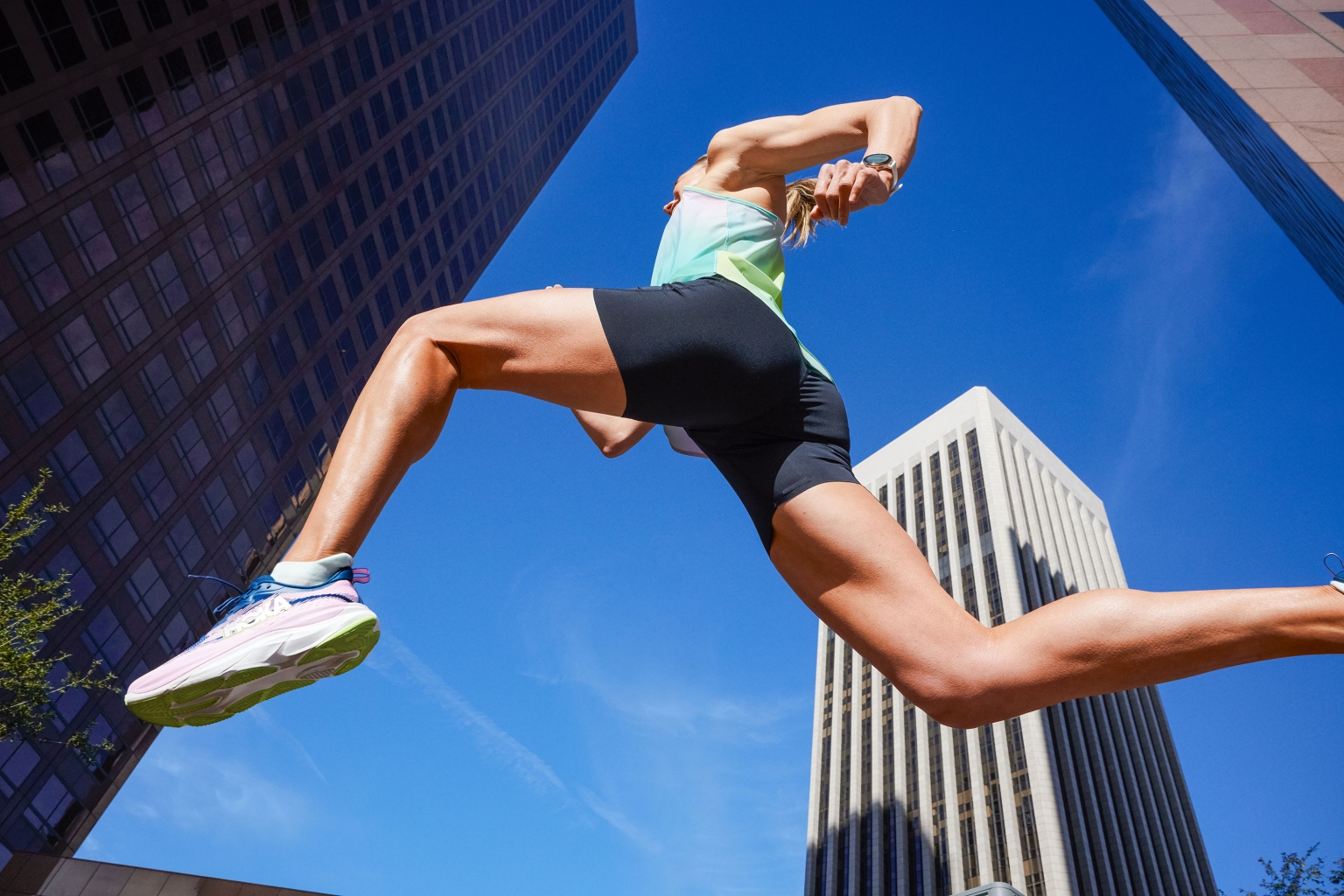 Woman in athletic wear leaps mid-air with high-rise buildings in an urban setting under a clear blue sky, showcasing fitness and urban athleticism.