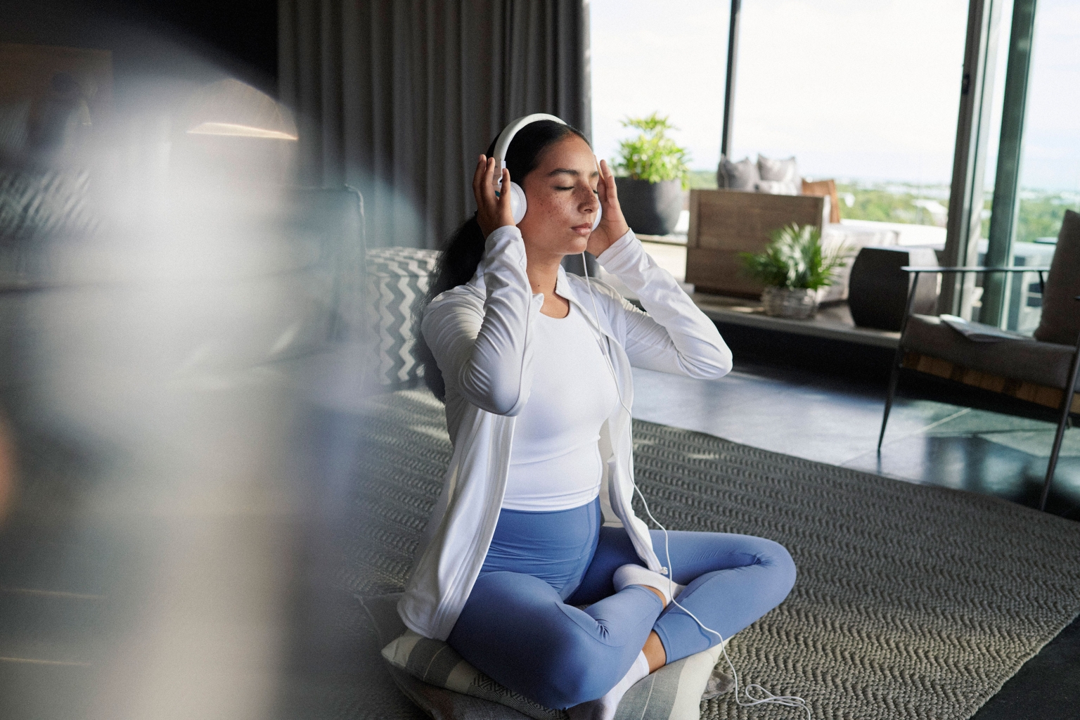 Woman in athleisure sits cross-legged on pillows indoors listening to headphones. She is in a room with neutral tones and large windows.