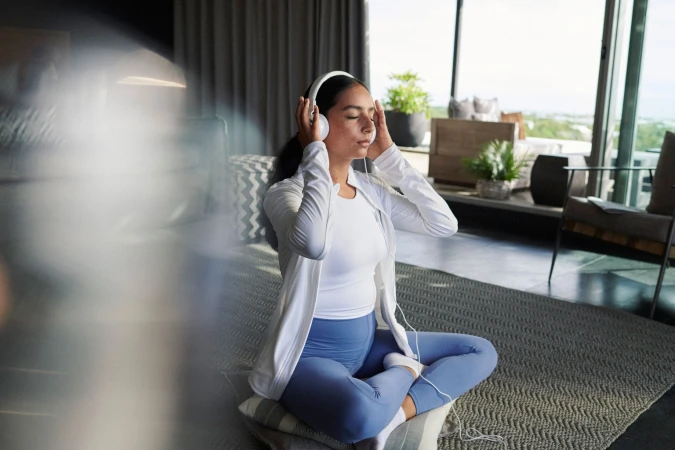 Woman in athleisure sits cross-legged on pillows indoors listening to headphones. She is in a room with neutral tones and large windows.