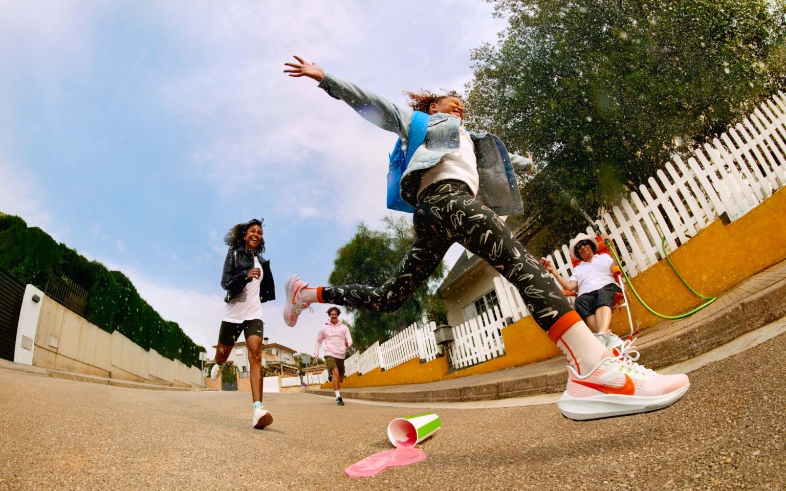 Excited young people run on a street, a child leaps in the air, sneakers visible, cup spills liquid. Outdoor shot with a sunny day sky.