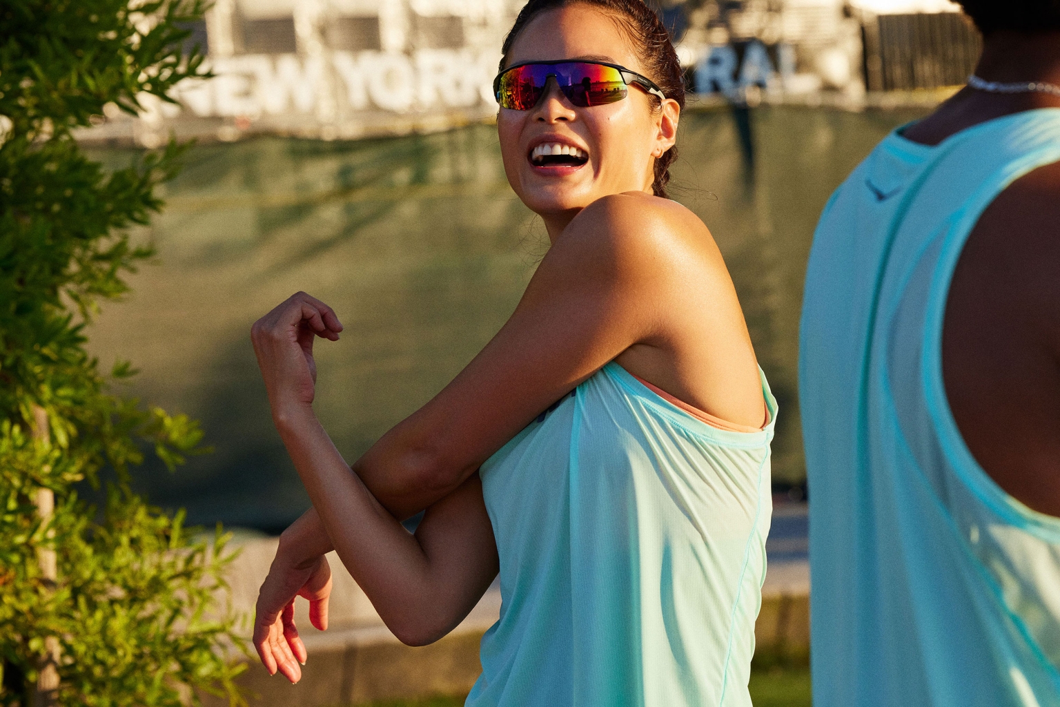 A woman in athletic wear stretches her arms outdoors while wearing sunglasses. Another person is partially visible in the frame.