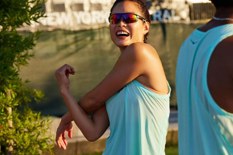 A woman in athletic wear stretches her arms outdoors while wearing sunglasses. Another person is partially visible in the frame.