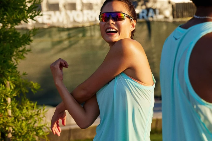 A woman in athletic wear stretches her arms outdoors while wearing sunglasses. Another person is partially visible in the frame.