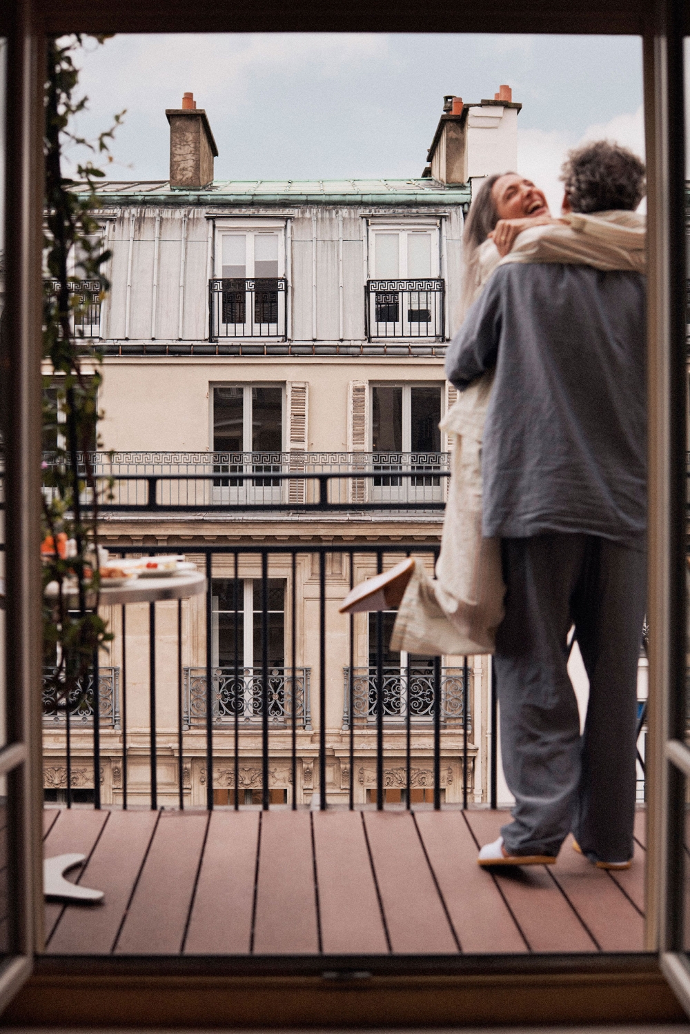 Affectionate couple embraces on a balcony with a view of Parisian buildings; she's smiling. Balcony has wood floors and a small table.