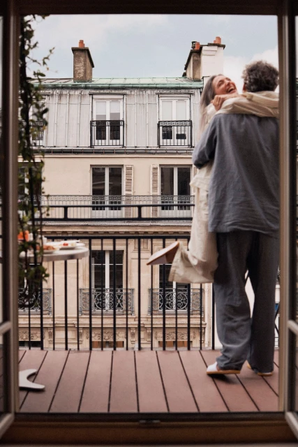 Affectionate couple embraces on a balcony with a view of Parisian buildings; she's smiling. Balcony has wood floors and a small table.